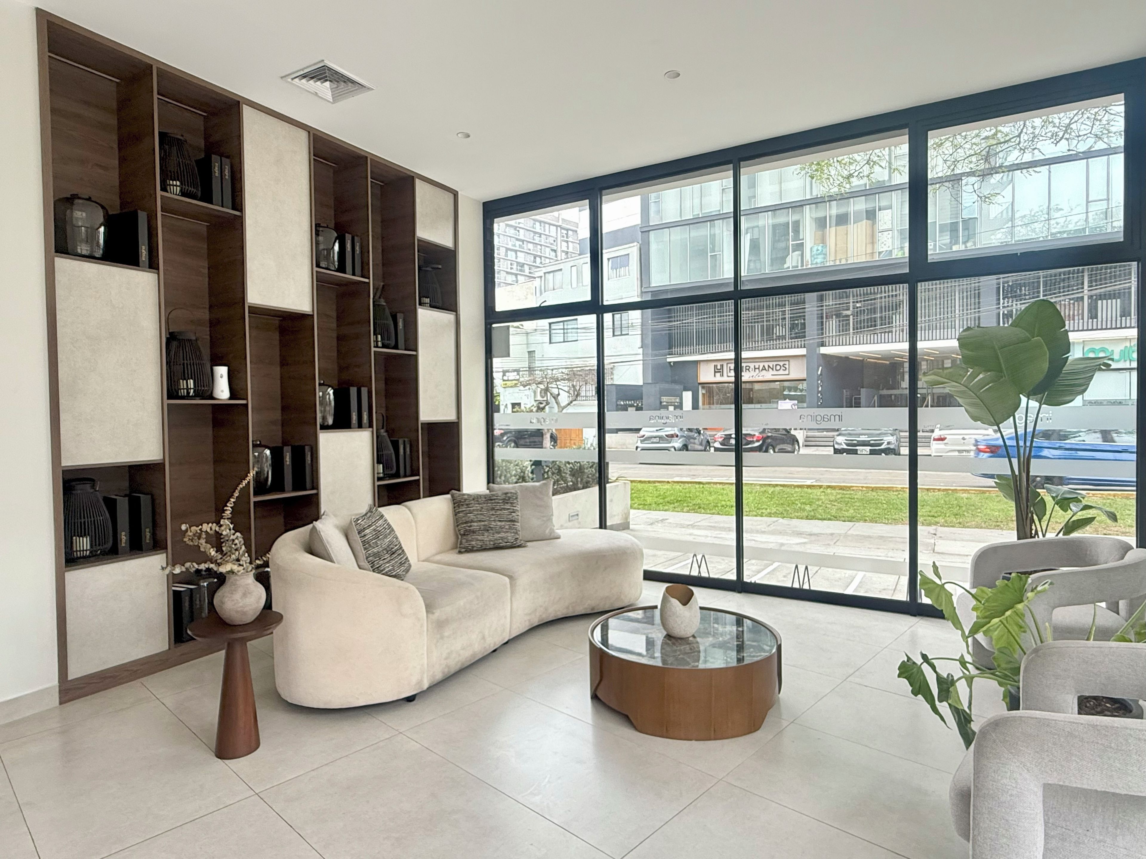 A modern living room with a large window, a sofa, a coffee table, and built-in shelving with various decorative items.