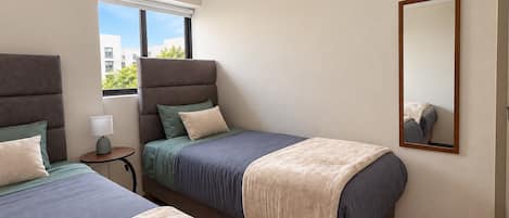 A hotel room with two beds, a bedside table, a lamp, a window with blinds, and a mirror on the wall.