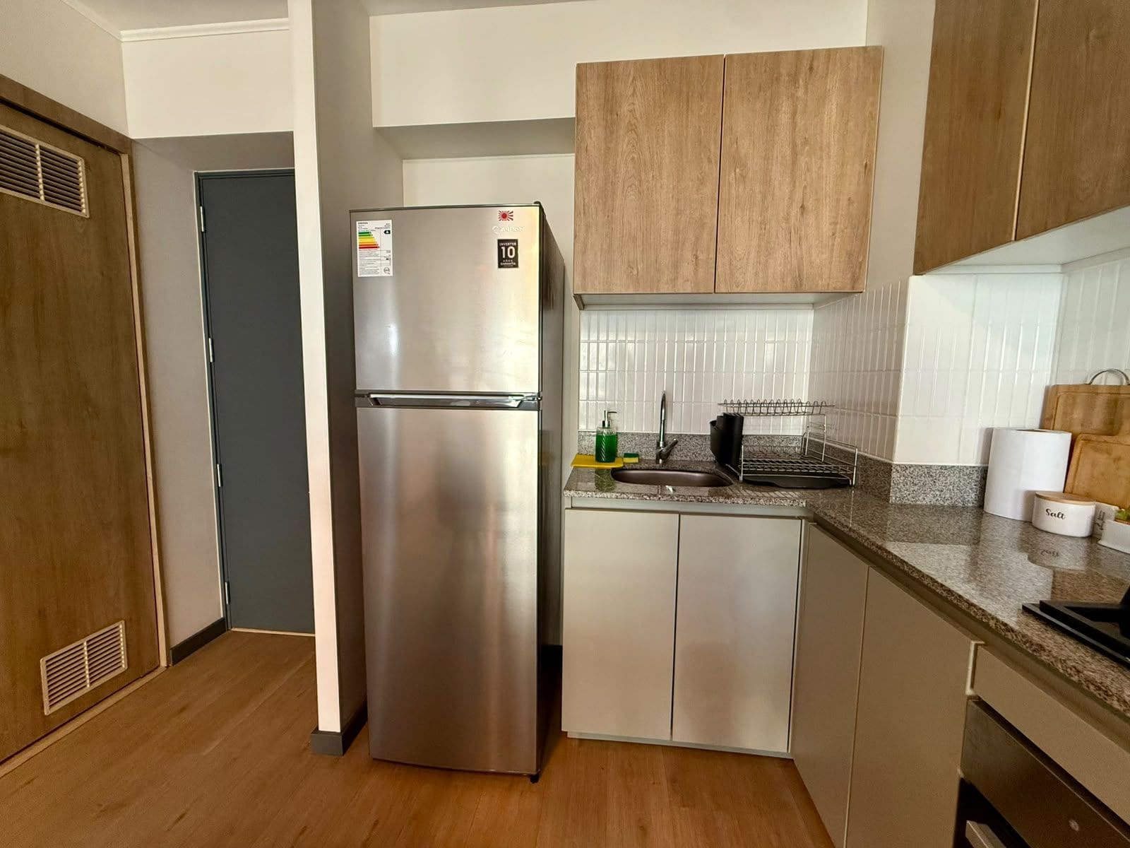 A compact kitchen with a stainless steel refrigerator, wooden cabinets, a granite countertop, and a built-in sink.
