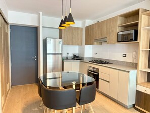 A modern kitchen with a glass dining table, black chairs, wooden cabinets, a stainless steel refrigerator, and a microwave on the countertop. - APT STYLE - MAGDALENA (Lima)