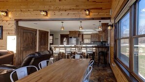 A rustic cabin interior with a wooden dining area, kitchen, and large windows.