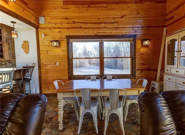 A wooden cabin interior with a dining table, chairs, and a large window.