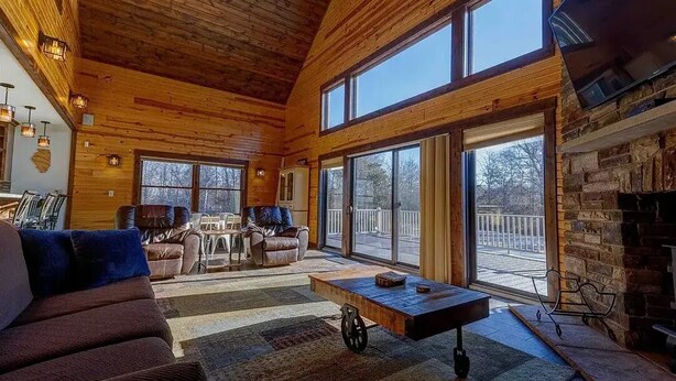 A spacious living room with a large sofa, a coffee table, and a fireplace. There are multiple windows providing natural light and a view of the outdoors.