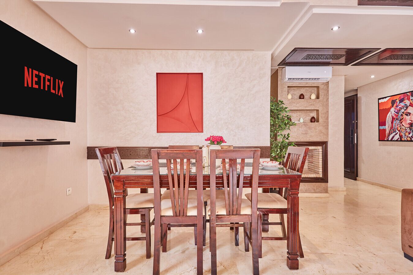 A dining area with a wooden table and chairs, a TV mounted on the wall, and a red abstract painting above the table.
