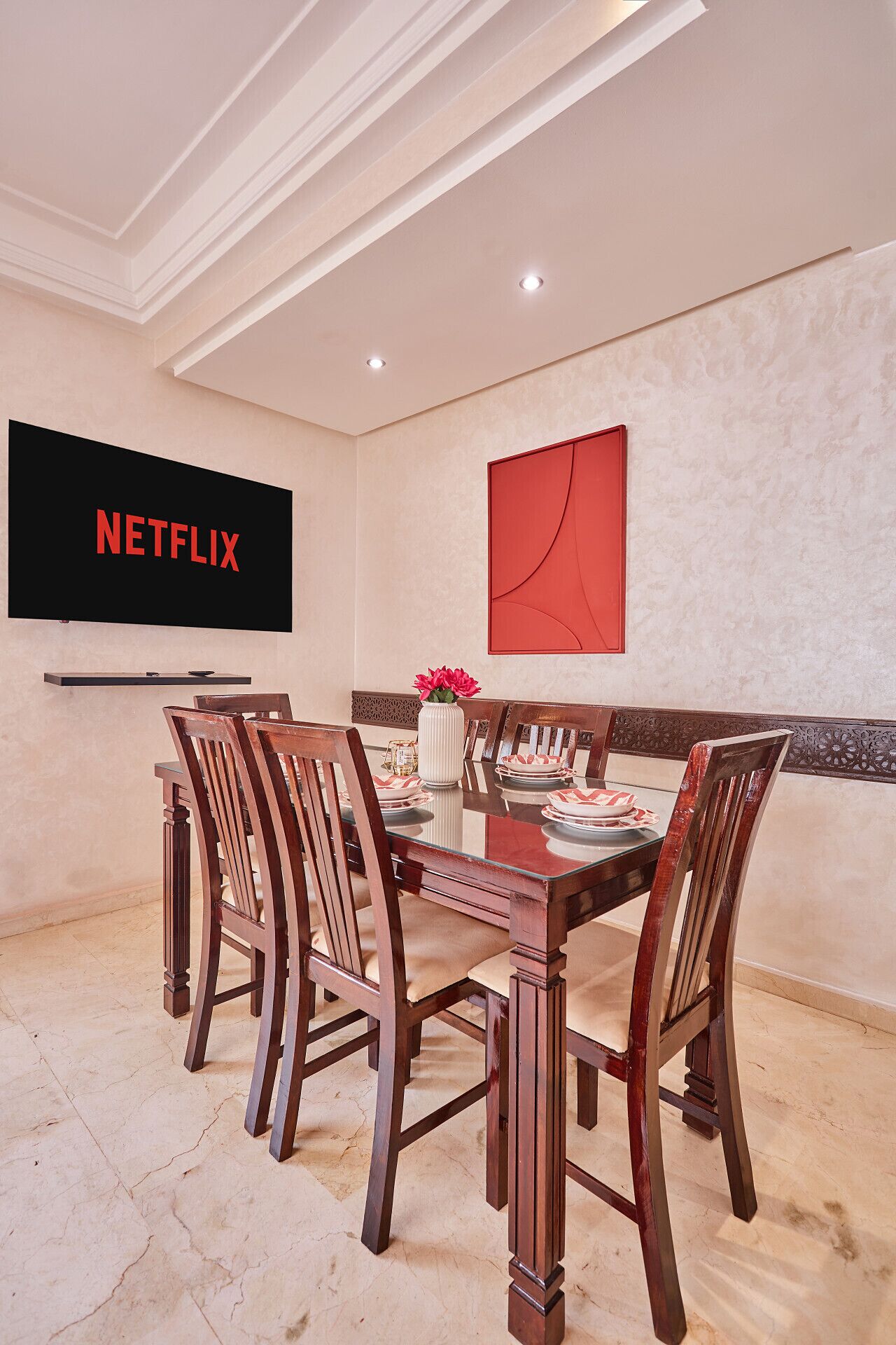 A dining area with a table set for four, a flat-screen TV mounted on the wall, and a decorative red artwork above the table.