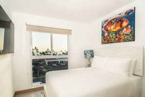 A hotel room with a large bed, a painting on the wall, a window with a view of a parking lot and palm trees, and a lamp on the nightstand. - Sun Style & Location Viva Jaco Condo (Jacó)