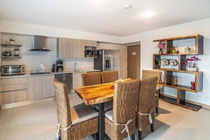 A modern kitchen with a dining area, featuring a wooden table and wicker chairs, stainless steel appliances, and a decorative shelf with plants. - Sun Style & Location Viva Jaco Condo (Jacó)