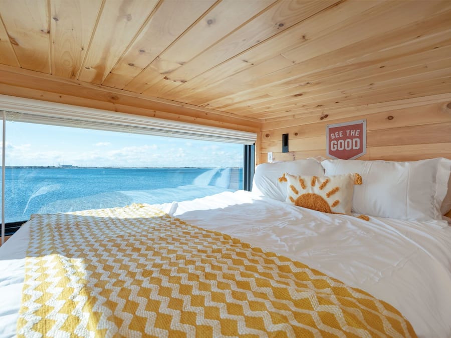 A wooden cabin bedroom with a bed, a view of the sea, and a sign that says "SEE THE GOOD".