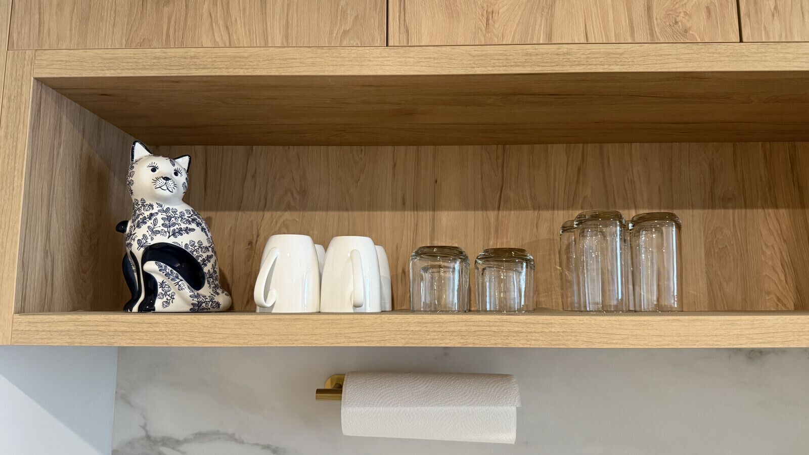 A wooden shelf with a ceramic cat figurine, white mugs, and clear glass jars.