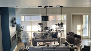 A modern living room with a grey sofa, a coffee table, and a dining table with a colorful bowl centerpiece. There are multiple hanging lights and a large window providing natural light. - Steps from Old Port. New, Beautifully Appointed Home. Sleeps up to 11. (Portland)