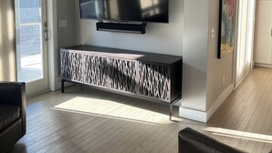 A modern living room with a flat-screen TV mounted on a wall, a patterned console table below, and a large window providing natural light. - Steps from Old Port. New, Beautifully Appointed Home. Sleeps up to 11. (Portland)