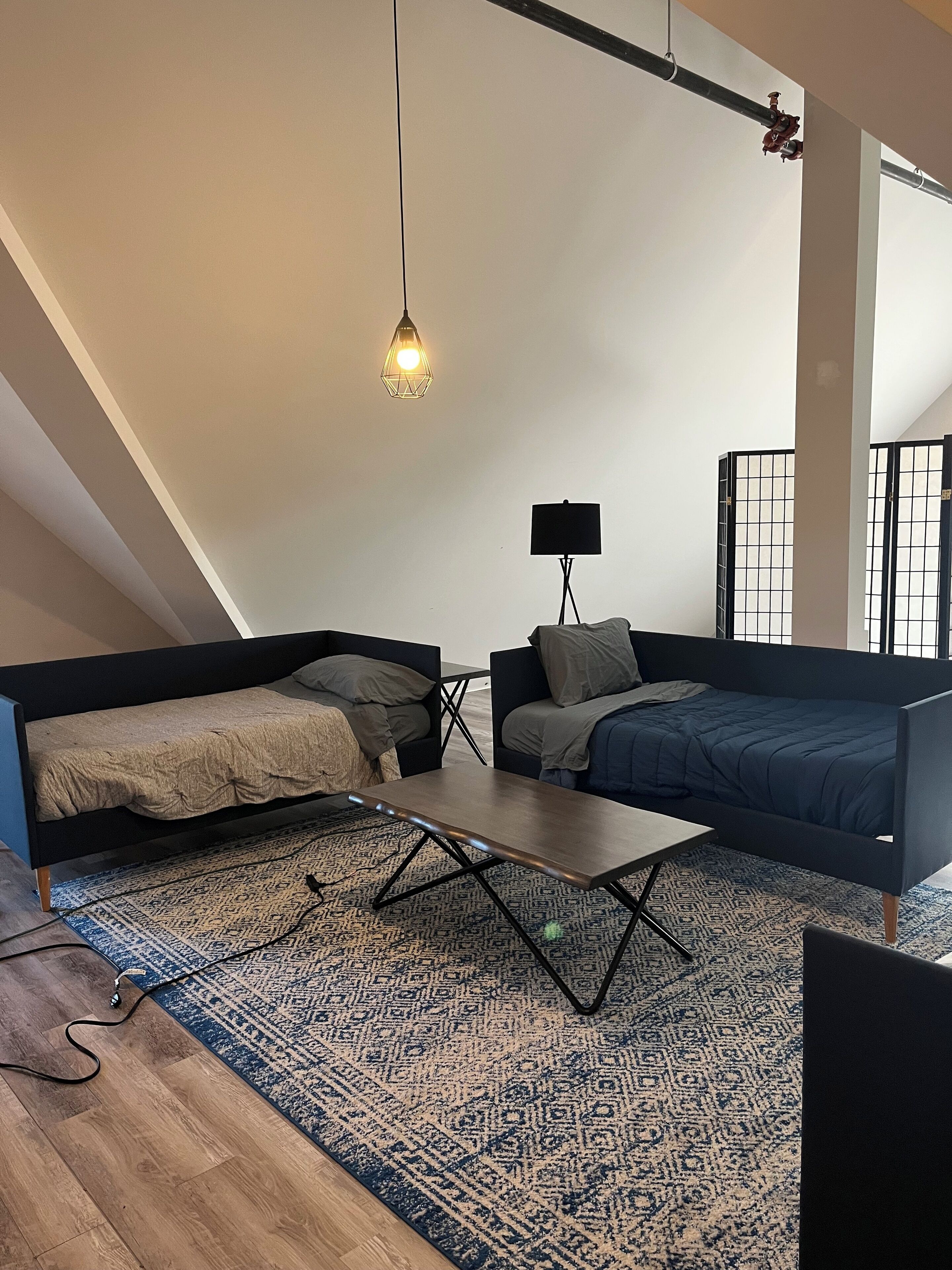 Un salon de style industriel, avec un haut plafond, des poutres apparentes, une grande fenêtre et un tapis à motifs.