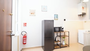 Une cuisine compacte avec un réfrigérateur noir, un four à micro-ondes et une machine à café placés sur une étagère en bois. Un extincteur est fixé au mur et il y a un lave-linge dans un coin.