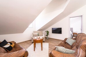 En modern lägenhet i loftstil med högt i tak, ett stort fönster, en platt-TV, lädersoffor och ett litet soffbord i trä.