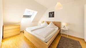 A bedroom with a wooden bed, a nightstand, a lamp, a framed picture, and a window with a view of a snowy landscape. - Ferienresidenz Hinterzarten FEWO 11 (Hinterzarten)