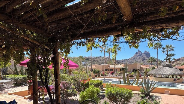 An outdoor patio with a table and chairs, surrounded by plants and a view of a pool and mountains.