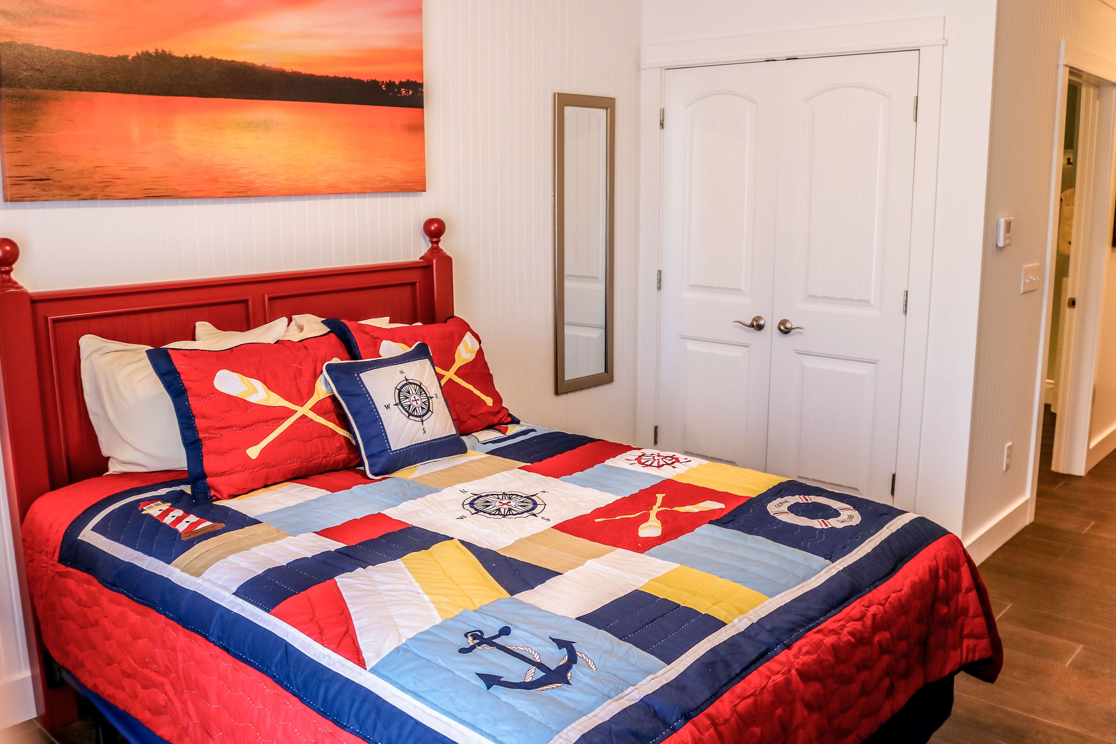 A bed with a nautical-themed quilt, a red headboard, and a painting of a sunset over water on the wall.
