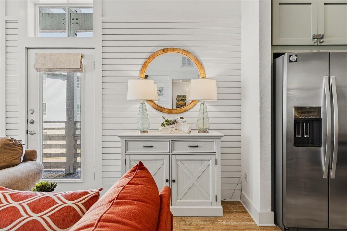Una habitación con pared de madera blanca, un espejo redondo con marco dorado, dos lámparas de pared, un aparador blanco, un refrigerador de acero inoxidable y un sofá rojo con estampado.