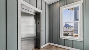 A room with a closed elevator, a window with a view of a house, and a herringbone wood floor. - Dnas Dunes TC 2202 (Port Aransas)