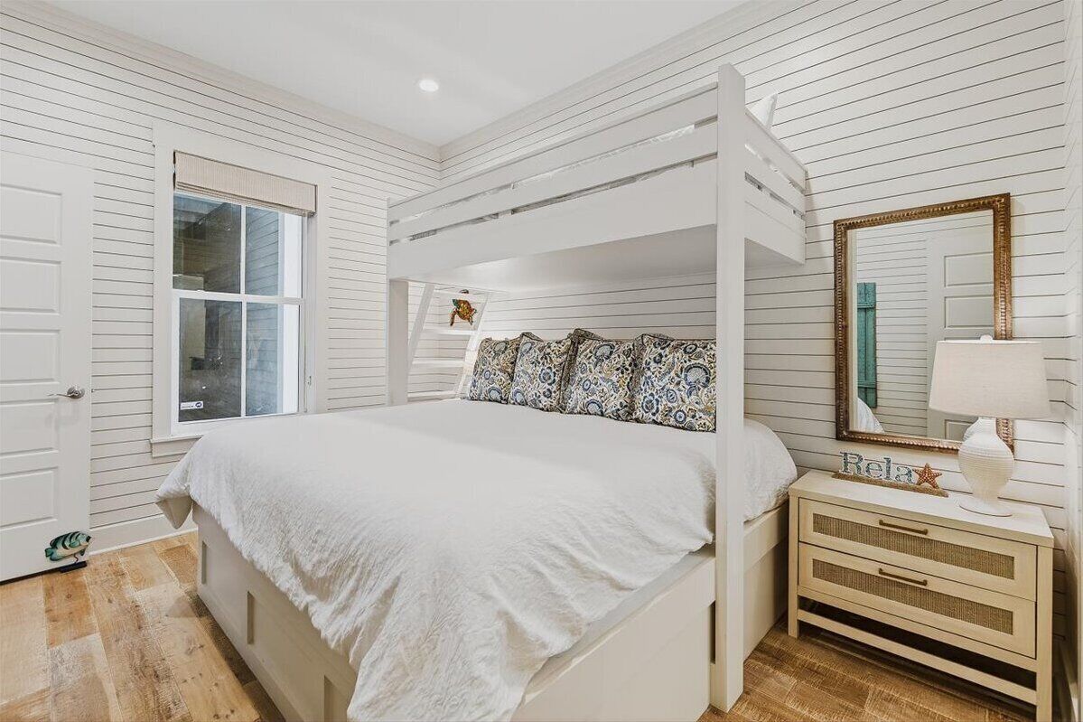 A bedroom with a bunk bed, a bedside table with a lamp, a mirror, and a window with blinds.