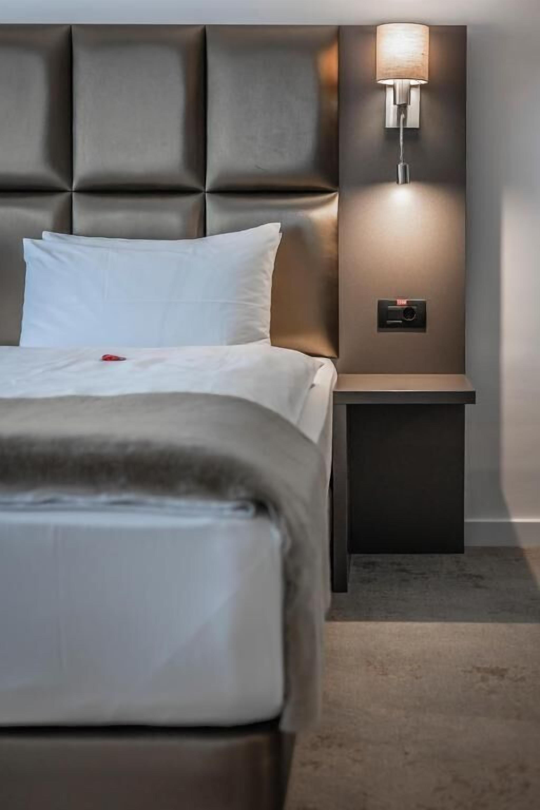 Ein modernes Hotelzimmer mit einem gepolsterten Kopfteil, einer Nachttischlampe und einem Bedienfeld auf dem Nachttisch.