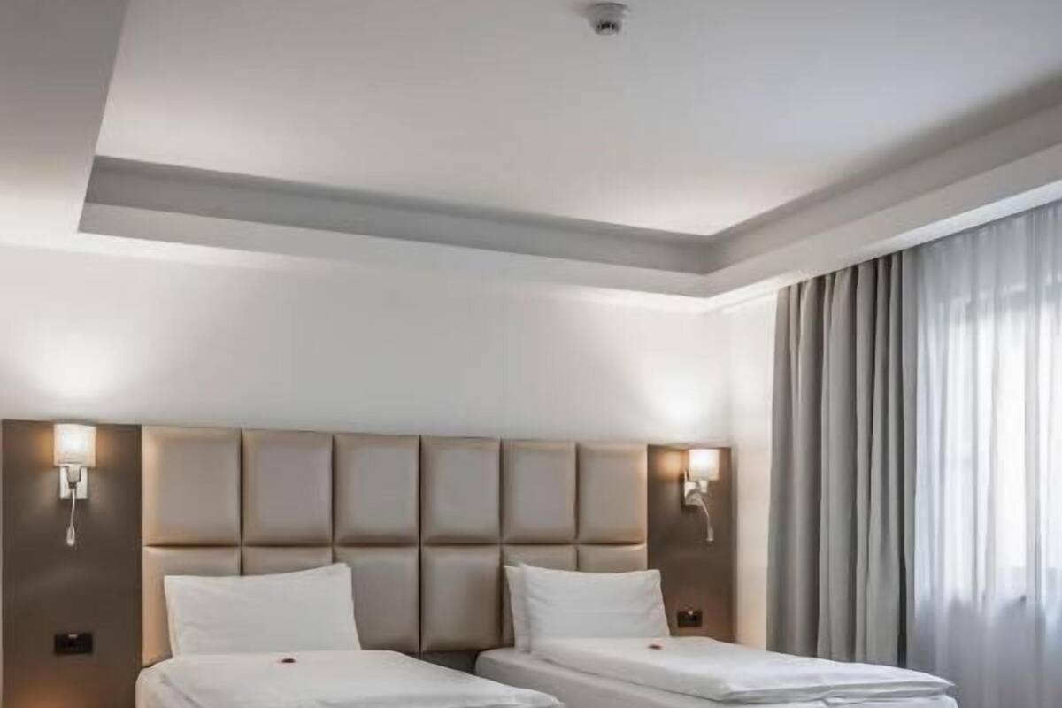 Hotellihuone, jossa on kaksi sänkyä, sängynpääty, yöpöytävalaisimet, pieni pöytä ja suuri ikkuna, jossa on verhot.