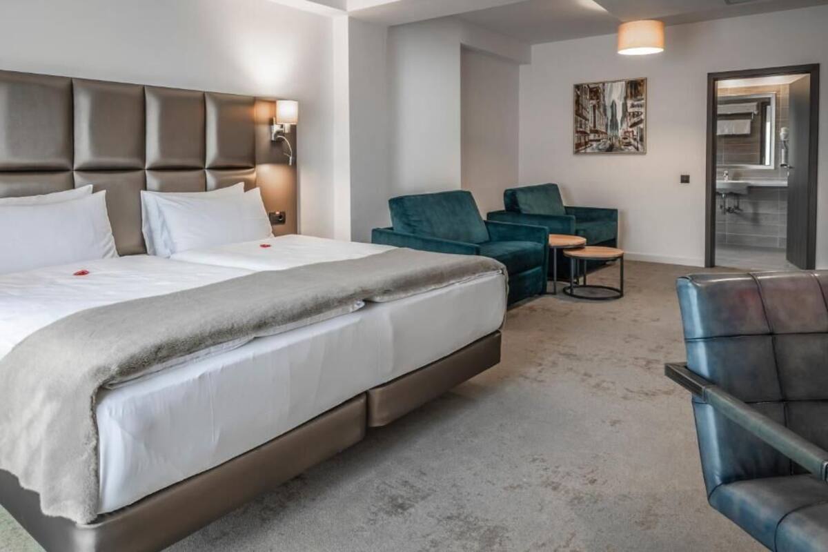 Hotellihuone, jossa on suuri sänky, kaksi tuolia, pieni pöytä ja kylpyhuone, joka näkyy avoimesta ovesta.