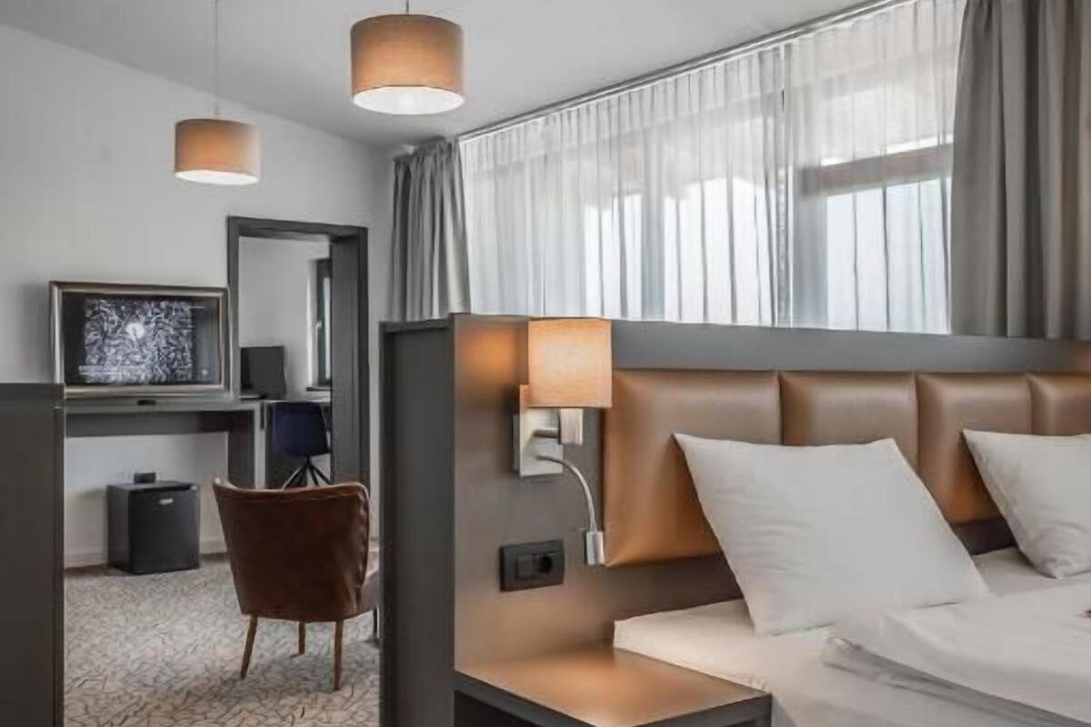 Moderni hotellihuone, jossa on sänky, yöpöydät, työpöytä tuolilla, televisio ja kuviollinen matto.