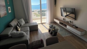 Un salon moderne comprenant un ensemble de canapés, une table basse, un téléviseur posé sur un meuble et une grande fenêtre donnant sur la mer.