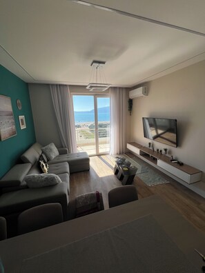 Un salon moderne comprenant un ensemble de canapés, une table basse, un téléviseur posé sur un meuble et une grande fenêtre donnant sur la mer.