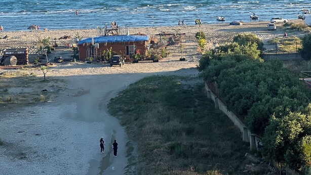 Une zone côtière avec une plage, un petit bâtiment doté d’un toit bleu, et une vue dégagée sur la mer et les montagnes lointaines.