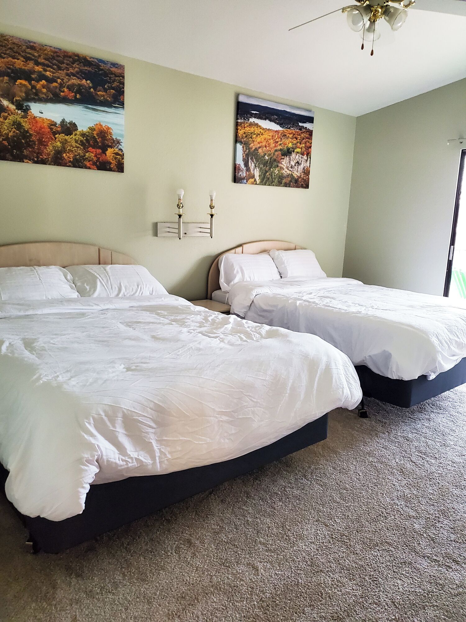 A hotel room with two beds, a ceiling fan, and two paintings of landscapes on the walls.