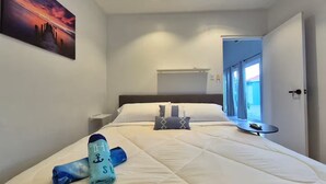Ein ordentlich bezogenes Bett mit einem zusammengerollten Handtuch und einer Decke, ein Nachttisch, ein Bild an der Wand und ein Fenster mit Vorhängen.