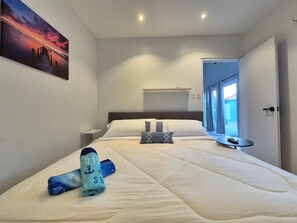 Ein ordentlich bezogenes Bett mit einem zusammengerollten Handtuch und einer Decke, ein Nachttisch, ein Bild an der Wand und ein Fenster mit Vorhängen.