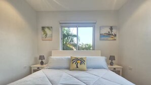 Un dormitorio con una cama grande, dos mesitas de noche, un ventanal con vistas a palmeras y dos cuadros colgados en la pared.