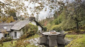 A rustic house with a stone chimney, a garden with a stone pathway, and wicker furniture around a central table. - Poppy Cottage - Coniston - Pass the Keys (Coniston)