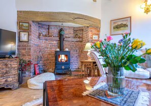 Ein gemütliches Wohnzimmer mit Kamin aus Backstein, einem Fernseher und einem Holztisch mit Blumen in einer Vase.
