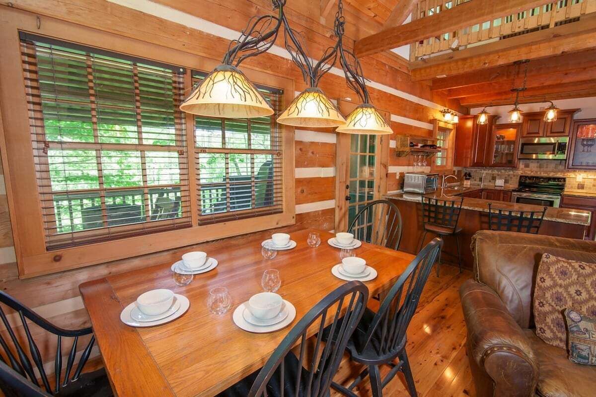 A wooden dining table set with white plates and glasses, surrounded by chairs, in a room with wooden walls and a kitchen area visible in the background.