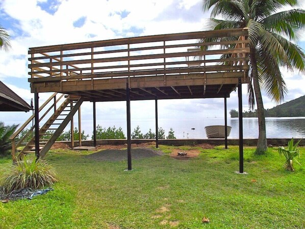 Una terraza de madera con una escalera que lleva a ella, rodeada de césped y palmeras, con vista a una masa de agua.