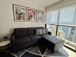 A modern living room with a dark sectional sofa, a glass coffee table, and a large window offering a city view. - APT VIZUALE - LINCE (Lima)