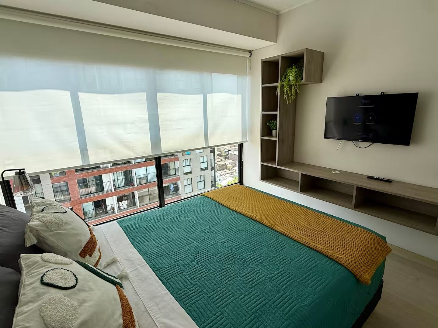 Un dormitorio moderno con un ventanal grande, una cama con colcha verde y amarilla, un televisor en la pared y una pequeña planta en un estante.