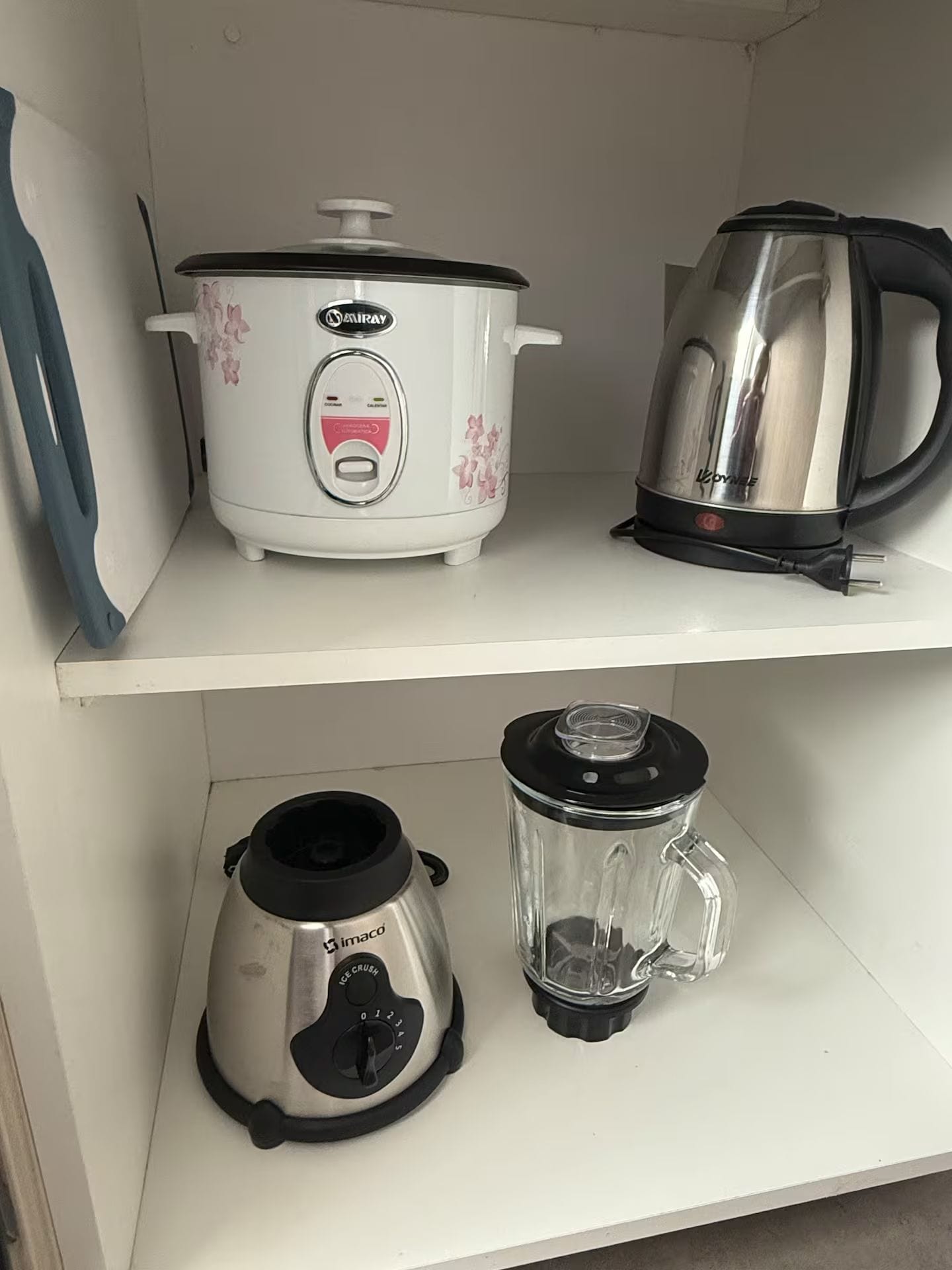 A kitchen shelf with a white slow cooker, a stainless steel electric kettle, and a black and silver food processor.