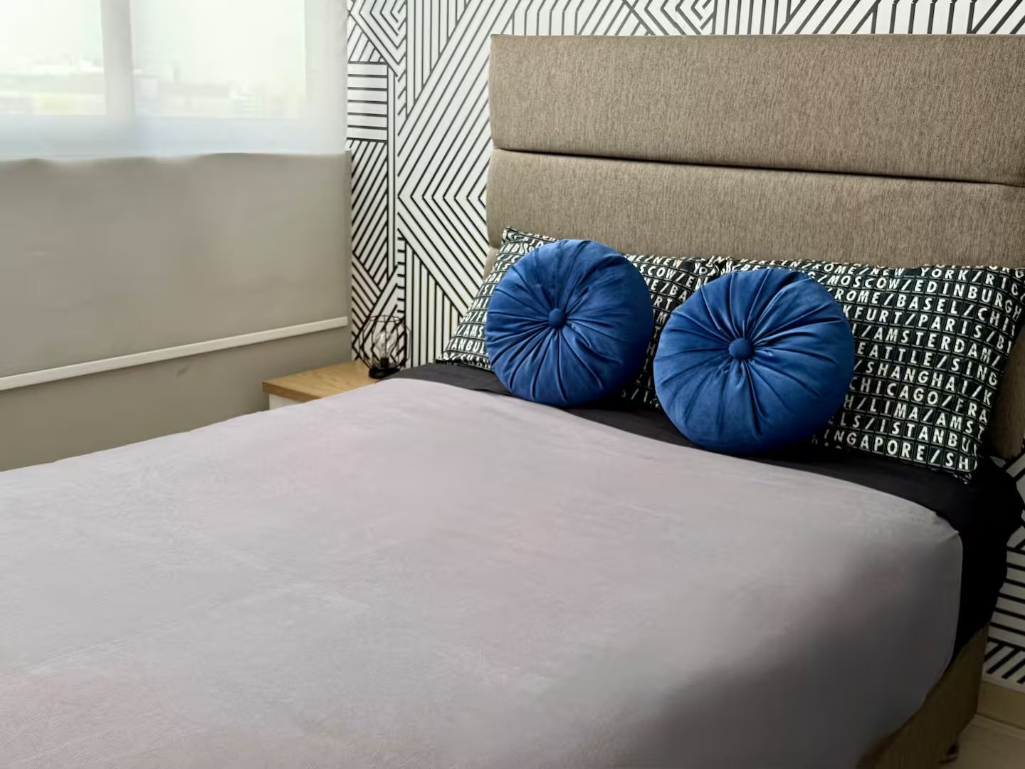 Una cama con colchón gris y dos almohadas azules, cabecera con diseño geométrico y mesita de noche con lámpara.