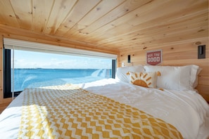Ein Schlafzimmer in einer Holzhütte mit einem Bett und Blick auf das Meer. Ein Schild trägt die Aufschrift 'SEE THE GOOD'.