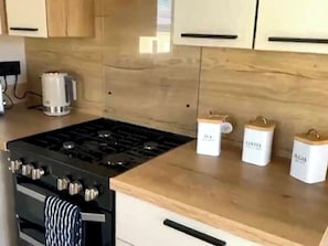 Eine Küche mit Gasherd, Backofen und Schränken. Auf der Arbeitsfläche stehen drei Behälter mit den Aufschriften 'Tee', 'Kaffee' und 'Zucker'.