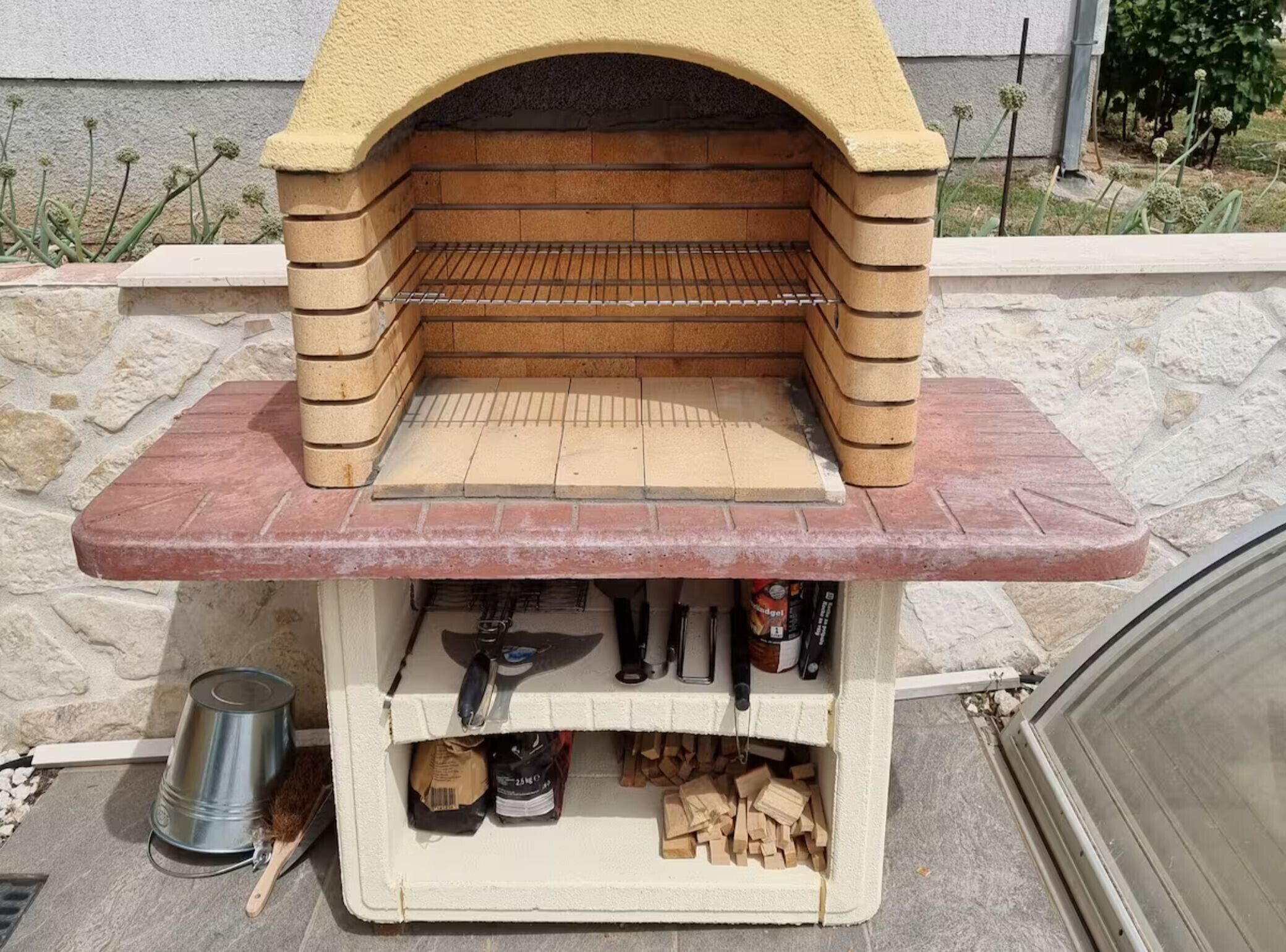 A stone-built outdoor pizza oven with a metal shelf and a brick base.
