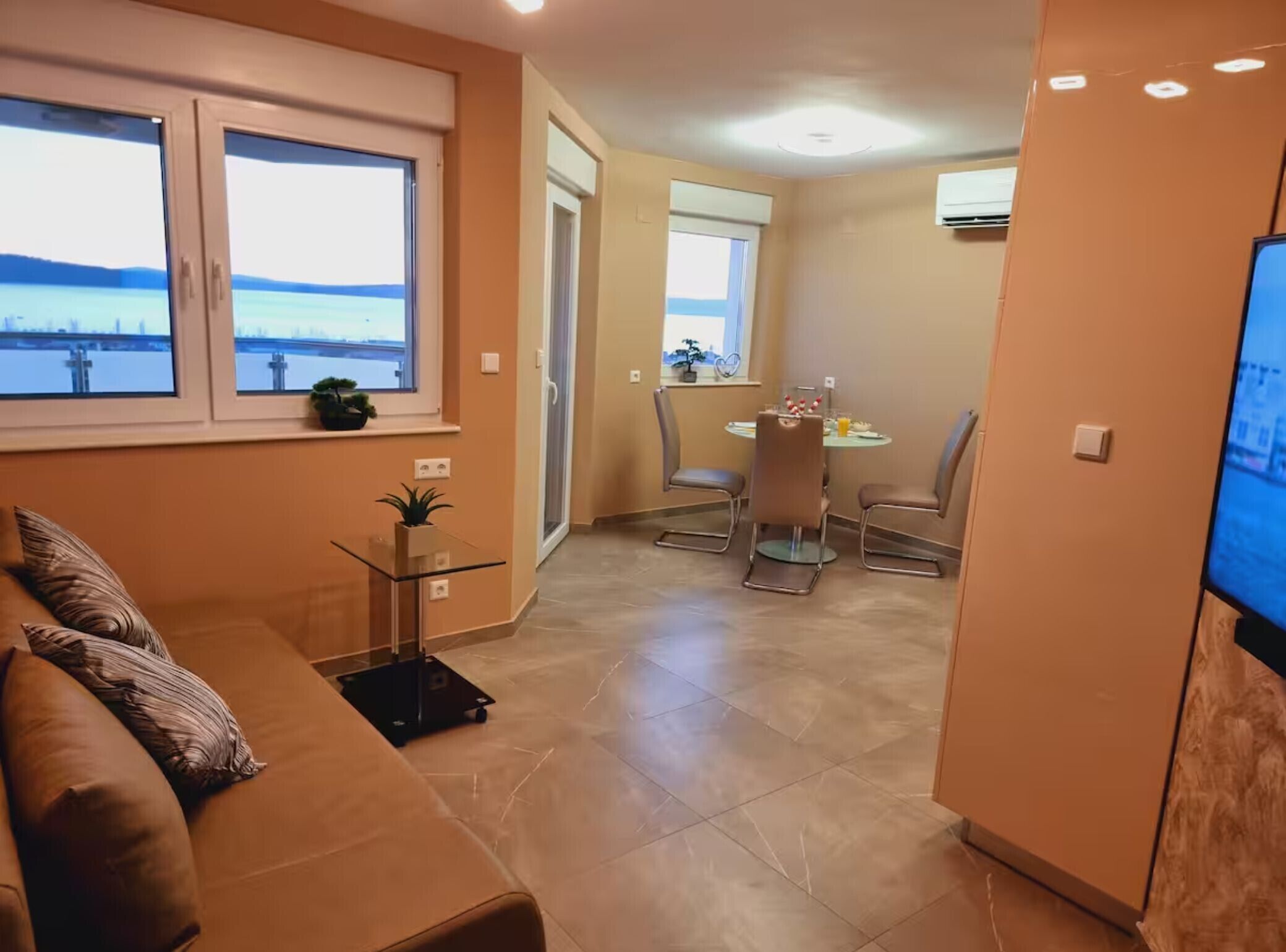 Un salon moderne avec un canapé marron, une table basse en verre et un téléviseur placé à droite. On y trouve également un coin repas avec une table et des chaises, et une vue sur la mer par les fenêtres.