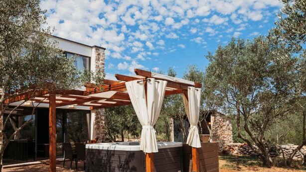 Ein Whirlpool unter einer Holzw pergola mit weißen Vorhängen, umgeben von Olivenbäumen und einer Steinmauer.