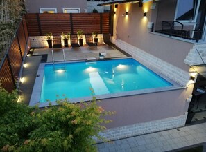 Ein rechteckiger Pool mit einer Holzterrasse und Liegestühlen, umgeben von einer Ziegelmauer und einem Holzzaun.