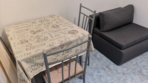 Una piccola stanza con un tavolo dal telaio in metallo ricoperto da una tovaglia con illustrazioni di cibi e bevande, un divano grigio e un pavimento piastrellato.